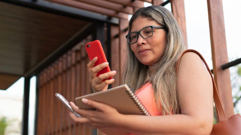 A middle-aged woman wearing glasses is holding a notepad and a pen in one hand and using a phone with the other hand.