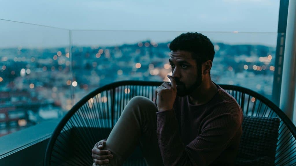 A depressed young African American man sitting on a balcony with an urban view in the evening.