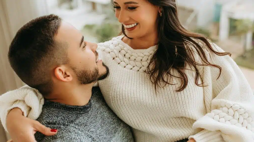 A smiling couple in sweaters sitting close and gazing at each other.
