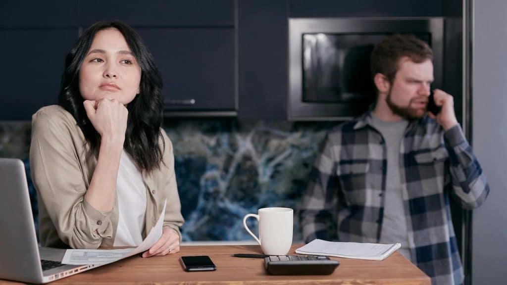 A woman looking worried at a table with bills while a man talks on the phone in the background.