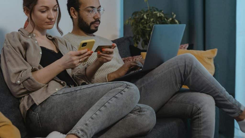 A couple sitting on a couch using smartphones and a laptop.