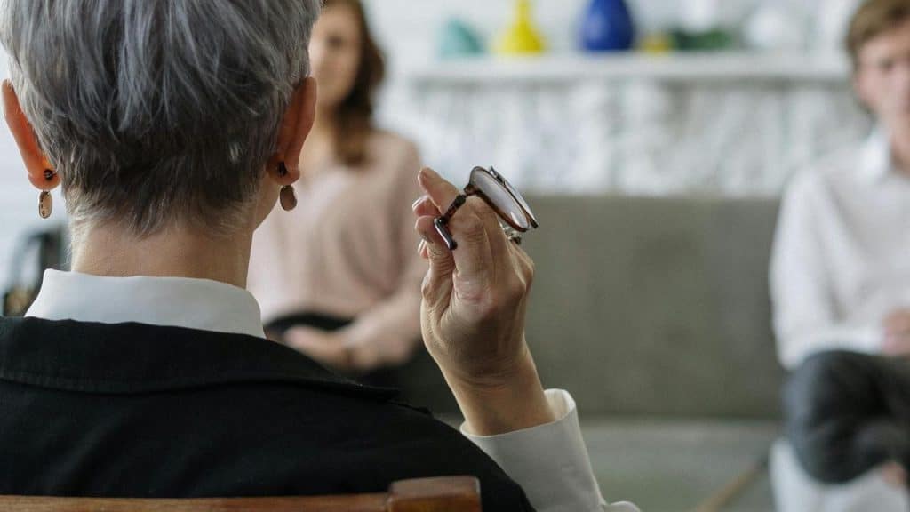 A therapist holding glasses while listening to two clients in a counseling session.