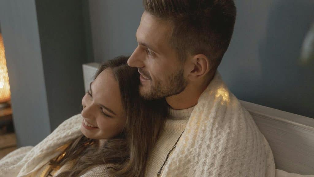 A couple cuddling under a blanket and smiling indoors.