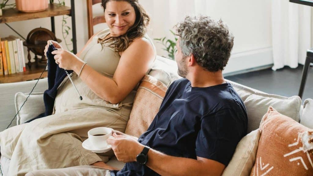A woman knitting on a couch while talking to a man holding a cup of coffee.
