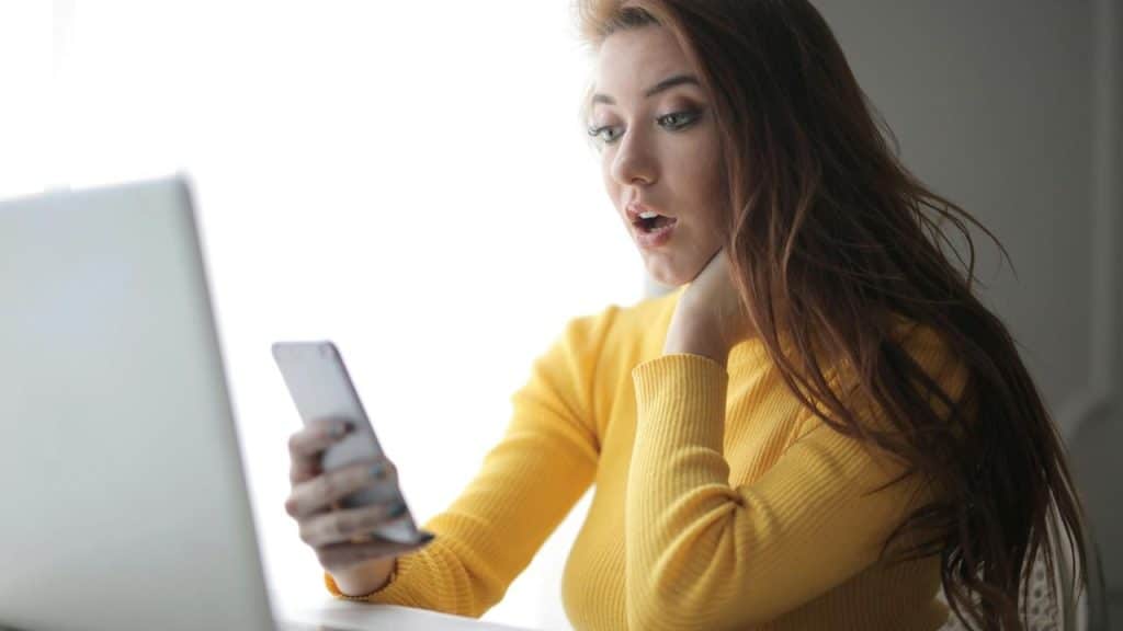A surprised woman in a yellow sweater looking at her phone beside a laptop.