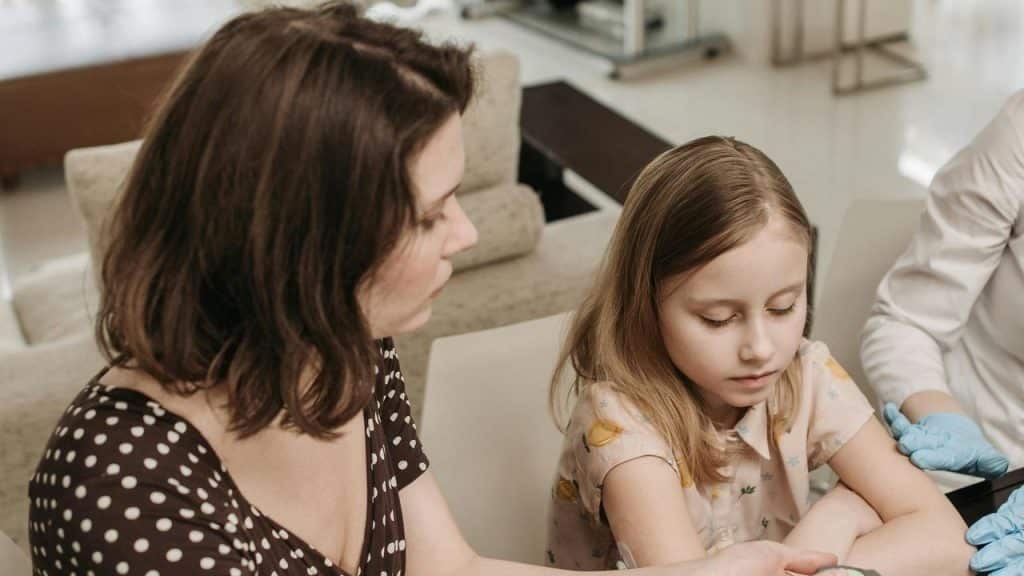 A mother sitting beside her daughter as a healthcare worker prepares to examine her arm.