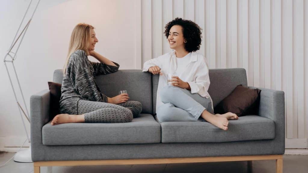 A pair of women sitting on a couch, smiling and talking while holding drinks.