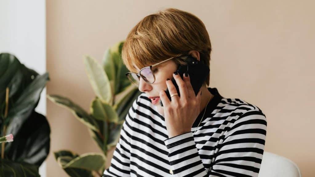 A woman with short hair and glasses talking on a smartphone indoors.
