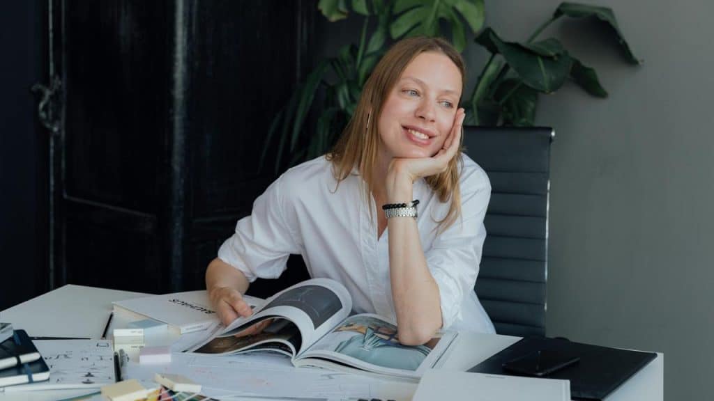 A woman smiling thoughtfully while reading a magazine at a desk.