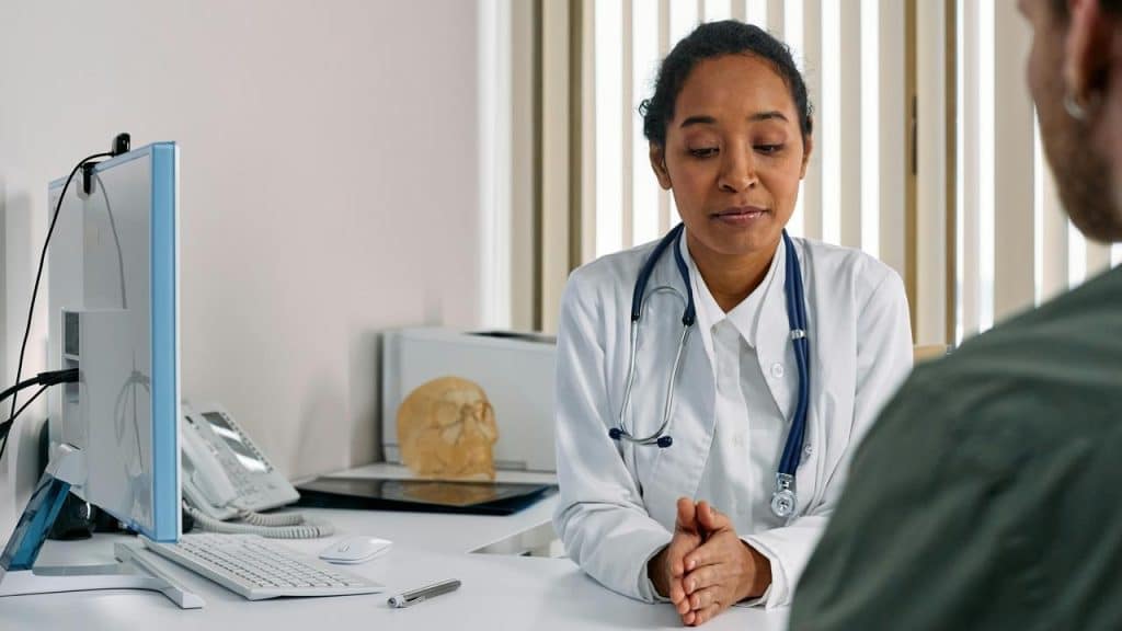 A doctor with a stethoscope sitting at a desk and speaking with a patient.