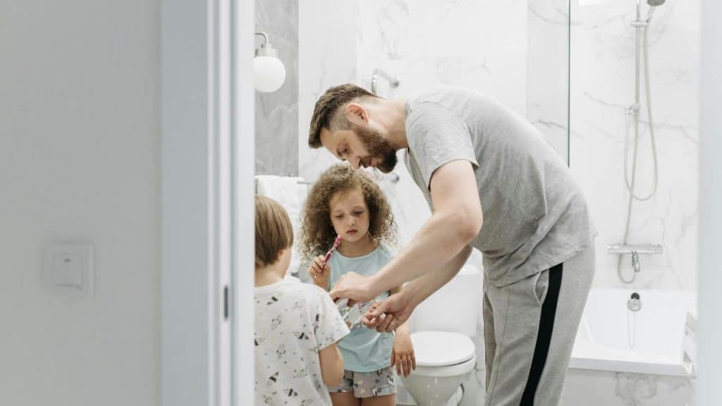 A father helping his two children brush their teeth in a bathroom.