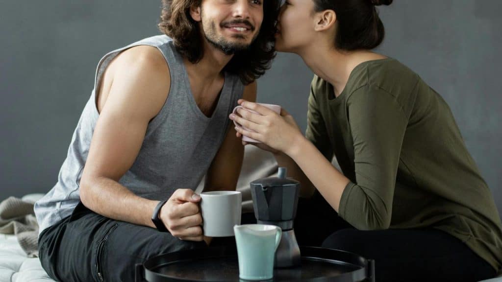 A woman whispering to a smiling man while they share coffee at a small table.