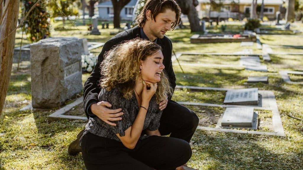 A grieving woman kneeling by a gravesite as a man comforts her in a cemetery.