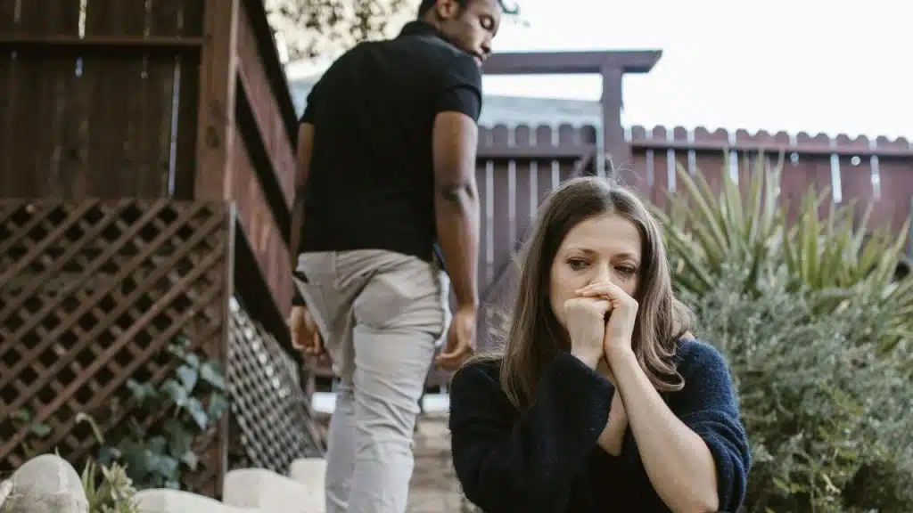 A distressed woman sitting outdoors while a man walks away in the background.
