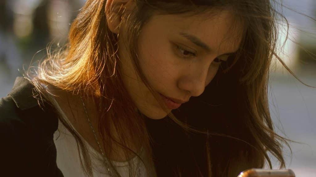 A young woman looking down thoughtfully at her phone in warm sunlight.