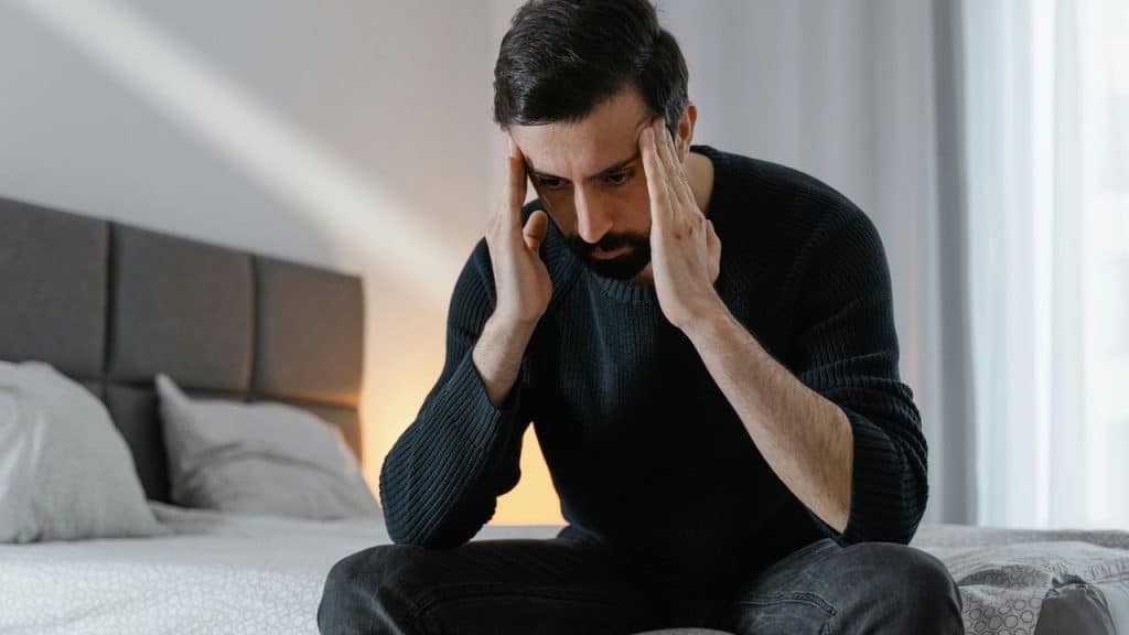A stressed man sitting on a bed with his hands pressed to his temples.