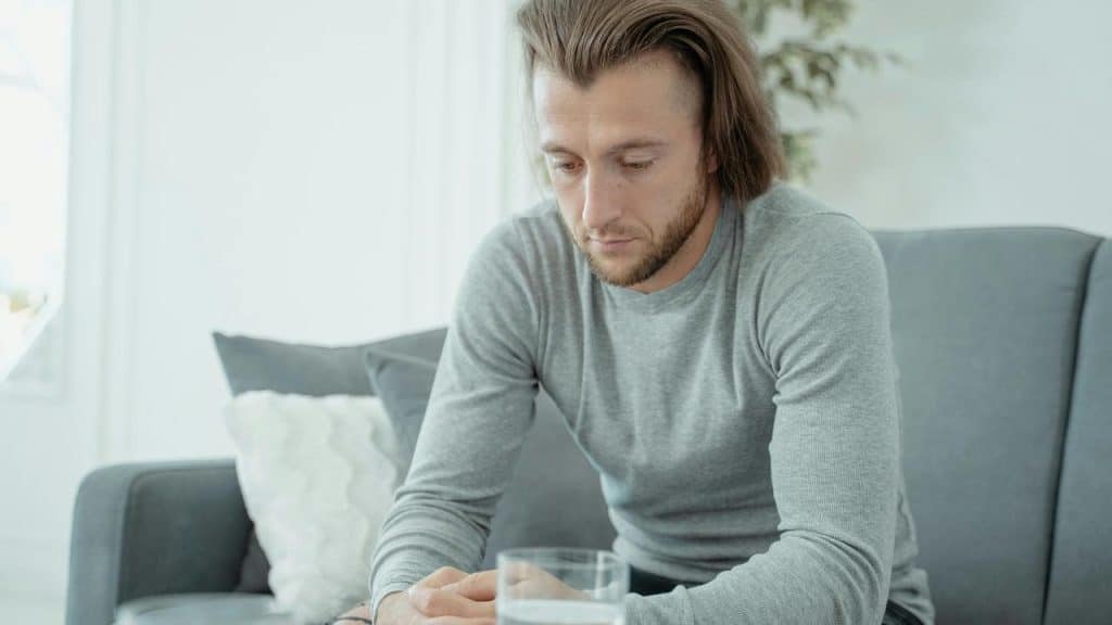 A somber man sitting on a couch with his hands clasped, looking down.