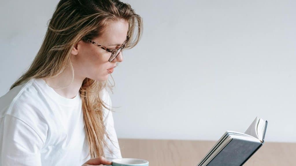 A woman wearing glasses reading a book while holding a cup.