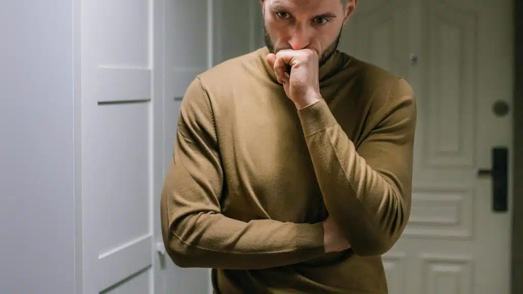 A thoughtful man in a brown sweater standing indoors with his hand covering his mouth.