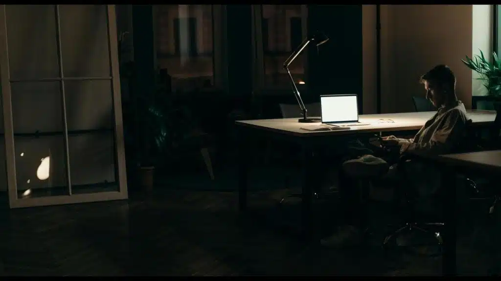 A man sitting at a desk at night, illuminated by a desk lamp and laptop screen.