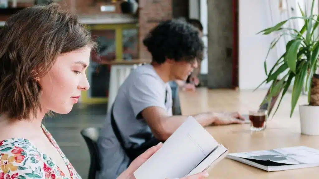 A woman reading a book at a table while a man works on a laptop in the background.