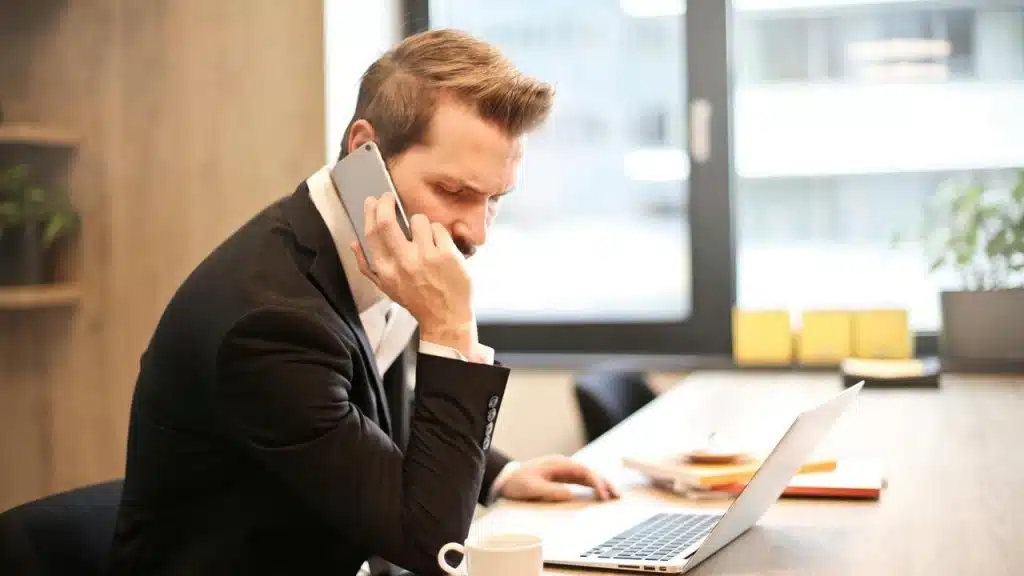A businessman talking on a smartphone while working on a laptop at his desk.