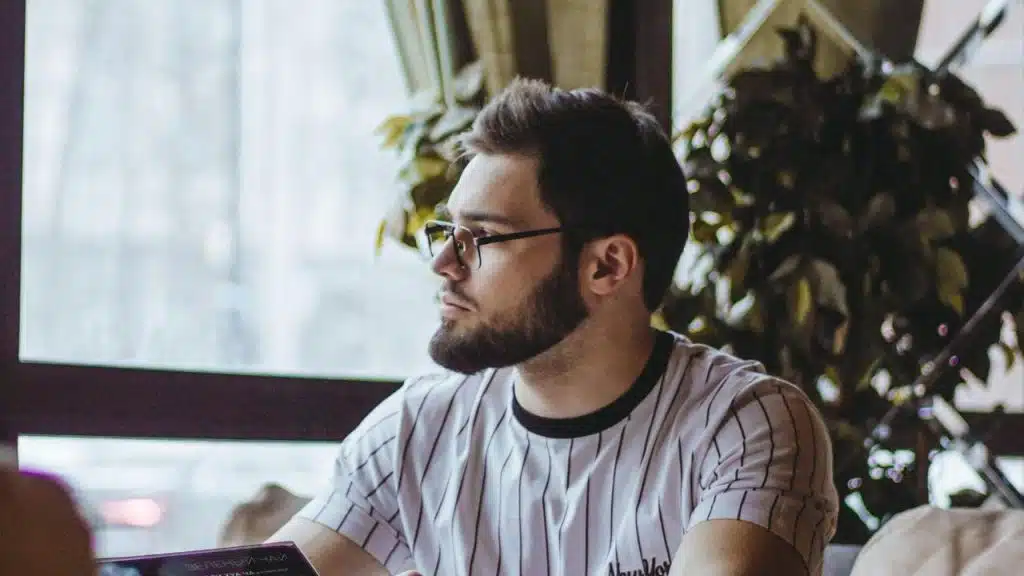 A man wearing glasses looking thoughtfully out a window indoors.