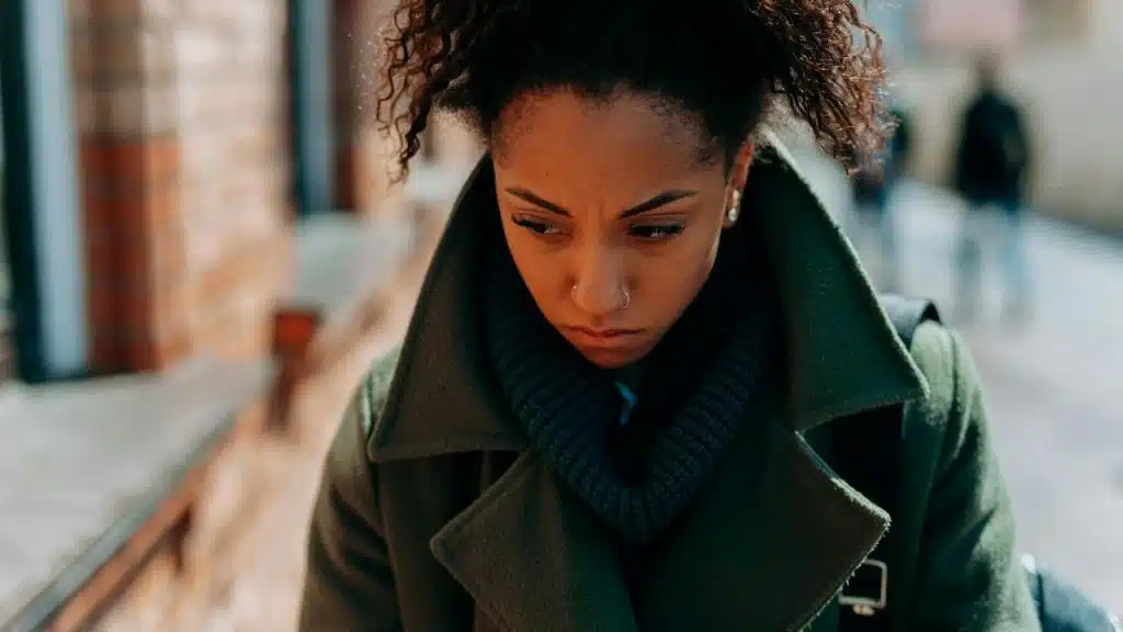 A woman in a green coat looking down with a worried expression on a city street.
