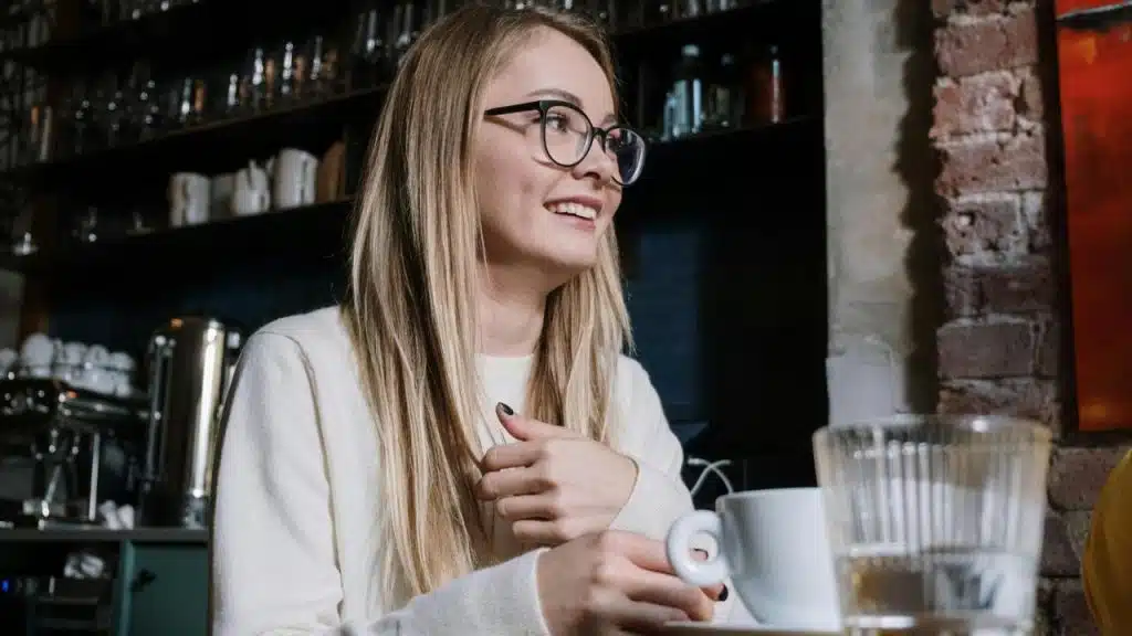 A smiling woman with glasses holds a coffee cup while sitting in a cozy café.