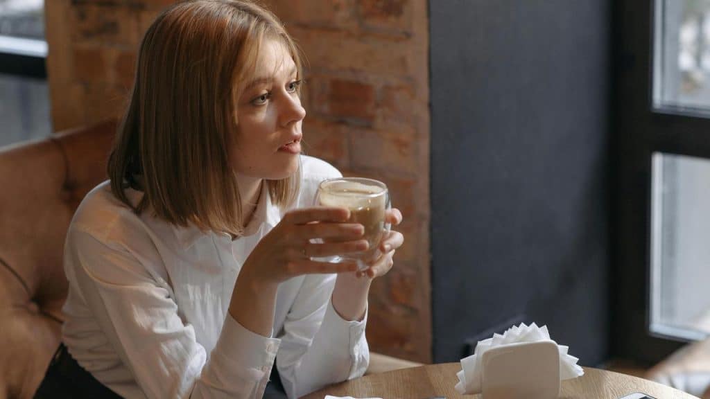 A woman sits by a window holding a glass cup of coffee in a cozy café.