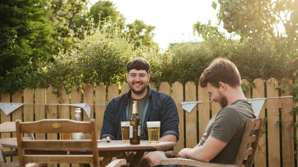 A pair of men sit at an outdoor table with beers in a sunny garden.