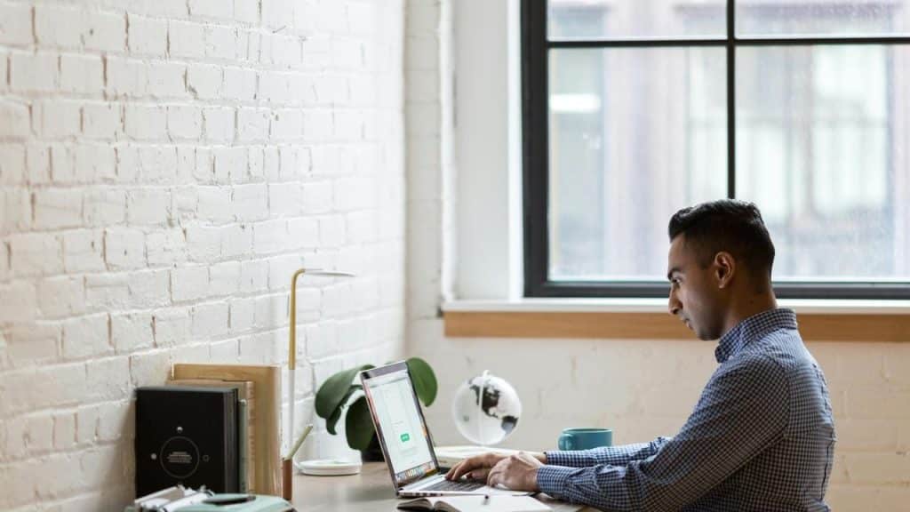 A man works on a laptop at a desk by a window.