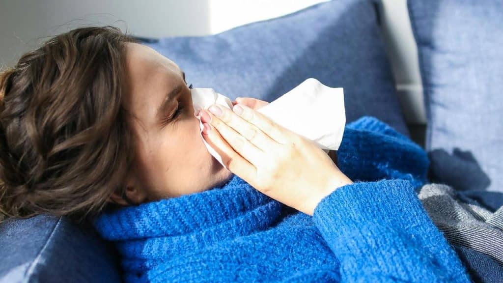 A woman lying on a couch blows her nose into a tissue.