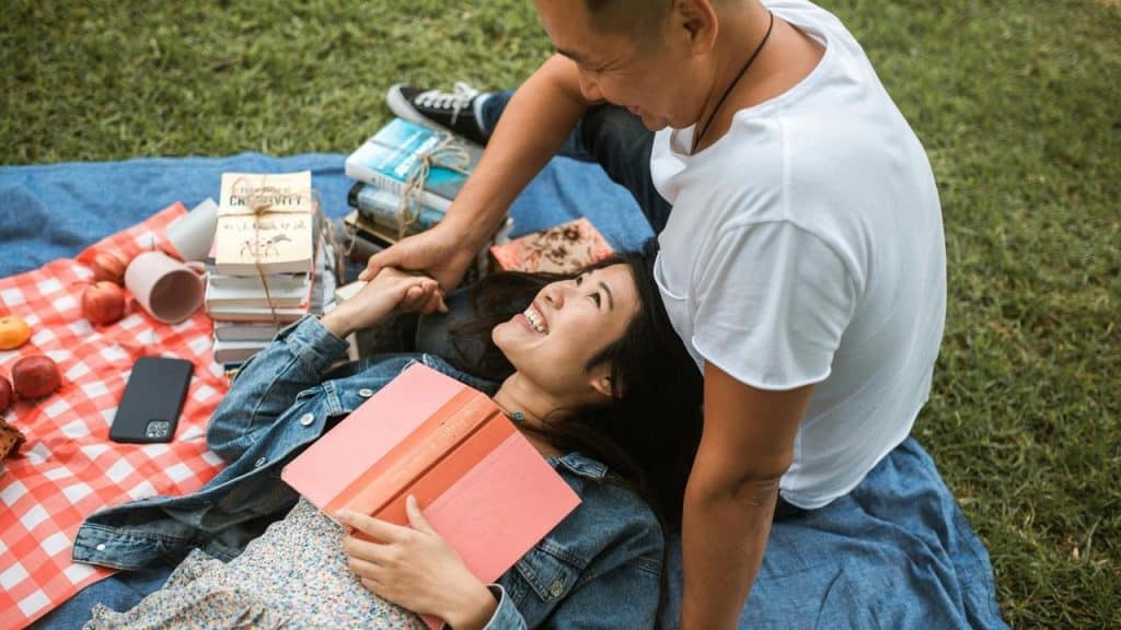 A couple relaxes on a picnic blanket, smiling and holding hands with books beside them.