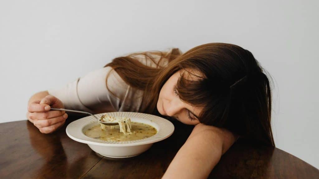A woman rests her head on a table beside a bowl of soup, holding a spoon.