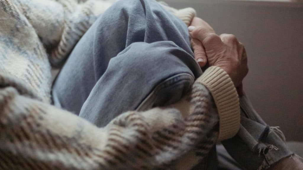 A person sits wrapped in a blanket with hands clasped around their knee.