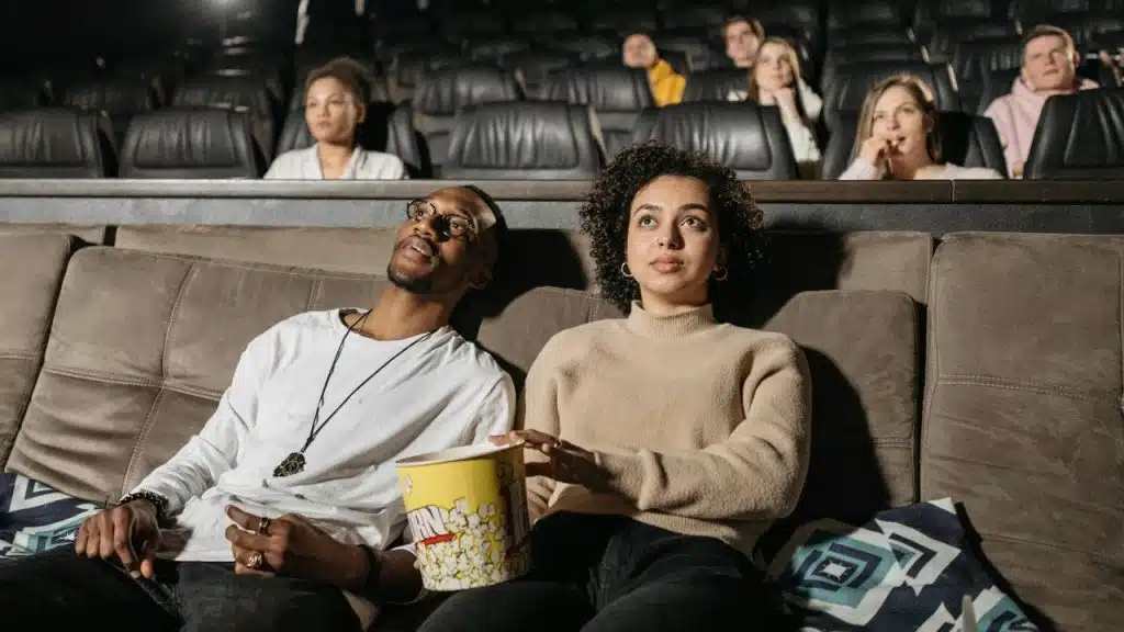 A couple sits in a movie theater watching a film with a bucket of popcorn.
