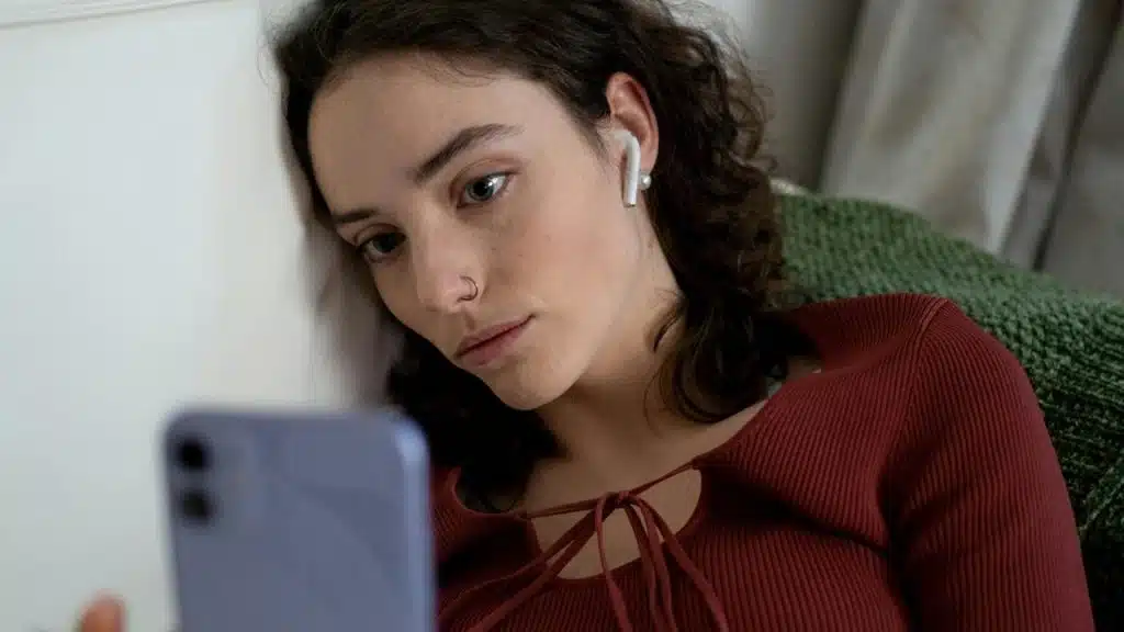 A woman with earbuds looks at her phone while resting against a wall.