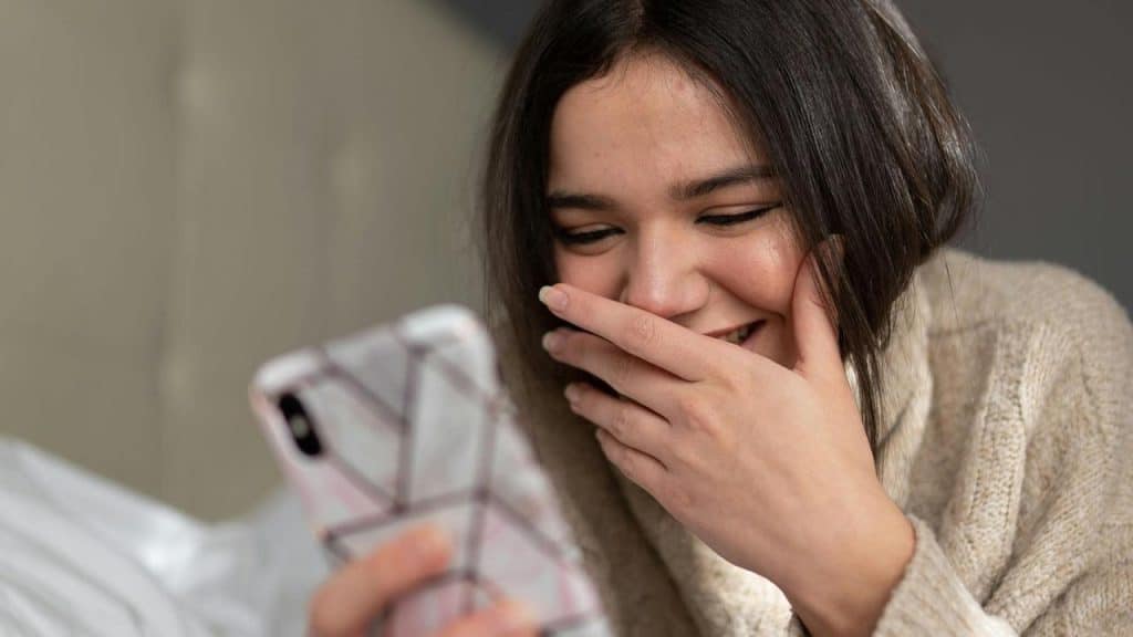 A woman smiles while looking at her phone and covering her mouth.