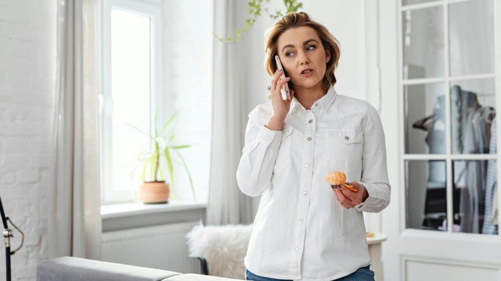 A woman talks on her phone while holding a peeled orange at home.