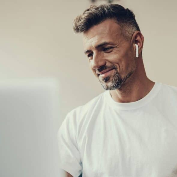 A smiling man wearing a white t-shirt and wireless earbuds looks down at a laptop.