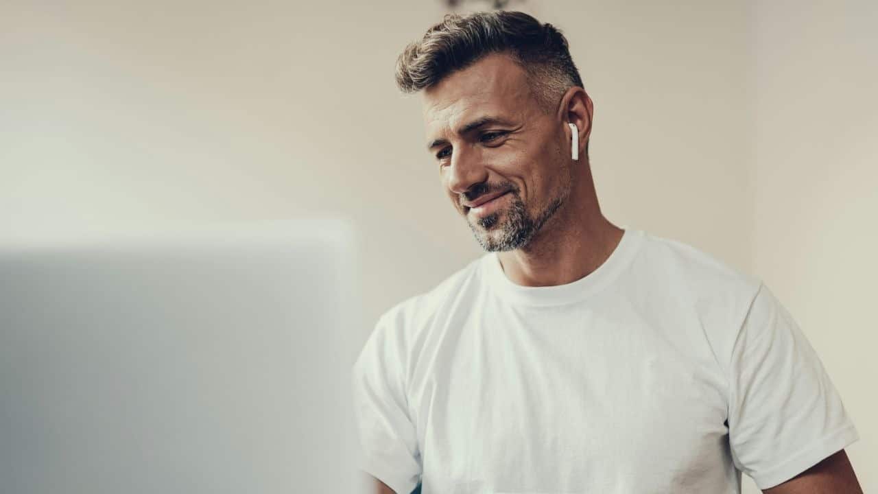 A smiling man wearing a white t-shirt and wireless earbuds looks down at a laptop.