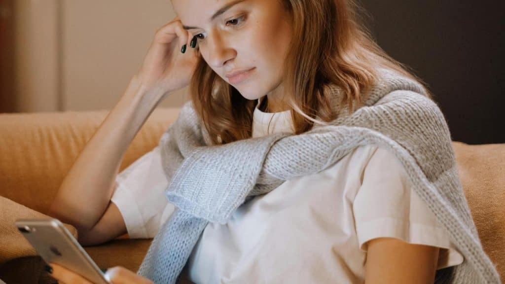 A woman sits on a couch looking at her phone with a thoughtful expression.