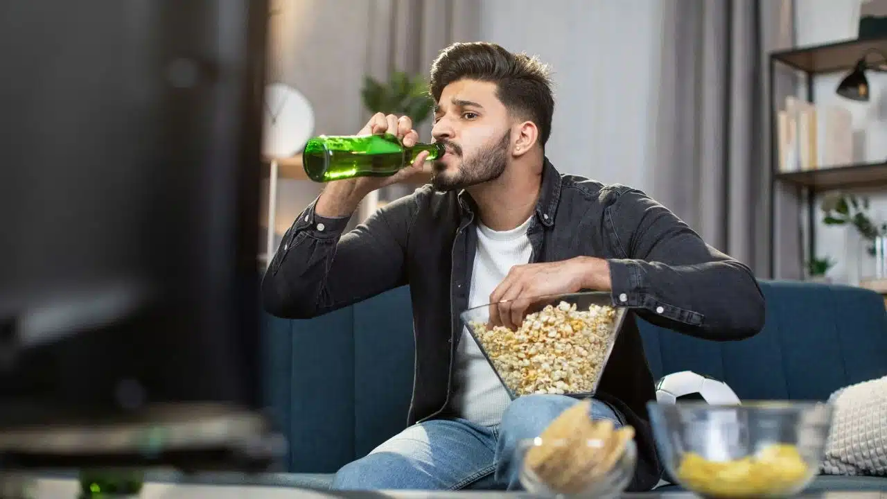 A man sits on a couch drinking from a green bottle while holding a popcorn bowl.