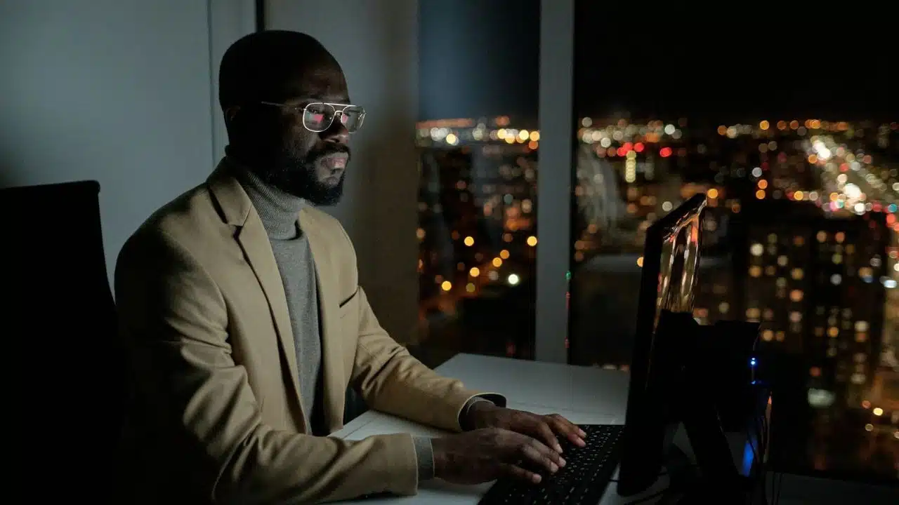 A man in a tan blazer and glasses types on a computer at night.