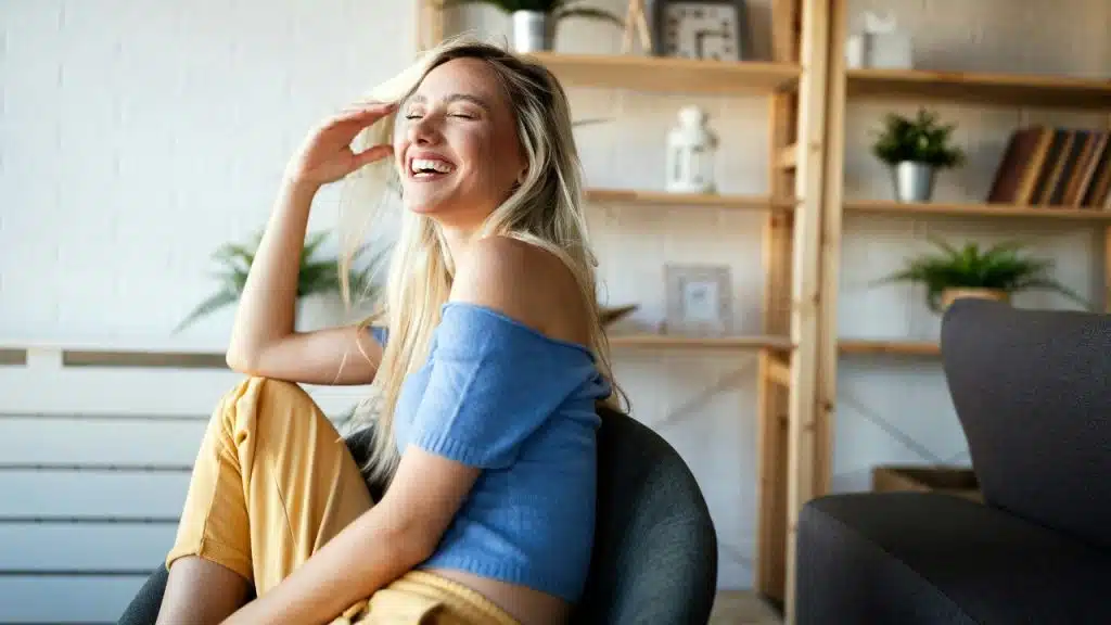 A blonde woman in a blue top laughs while sitting in a chair at home.