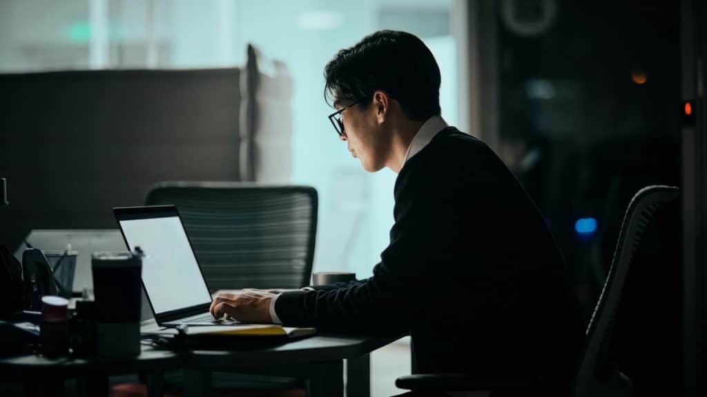 A man wearing glasses sits at a desk in a dark room working on a laptop.