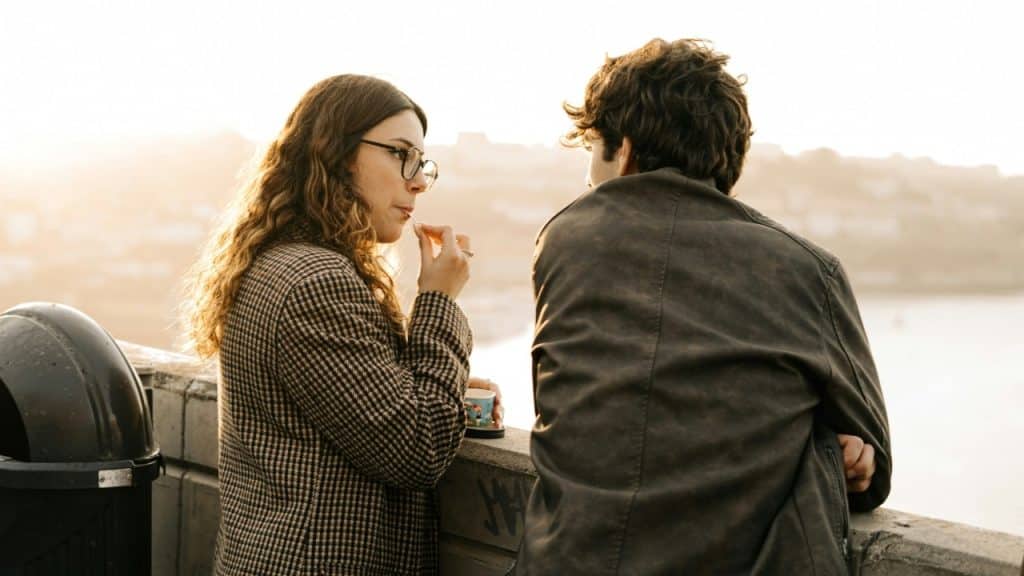A woman in a patterned coat eats while sitting on a wall next to a man.
