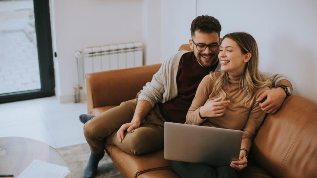 A man and woman sit on a brown leather couch looking at a laptop and laughing.