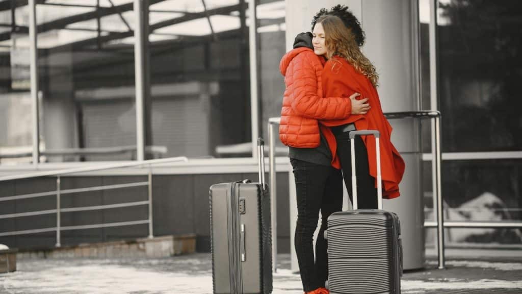 Two people embrace outdoors next to two large suitcases on a sidewalk with glass buildings.